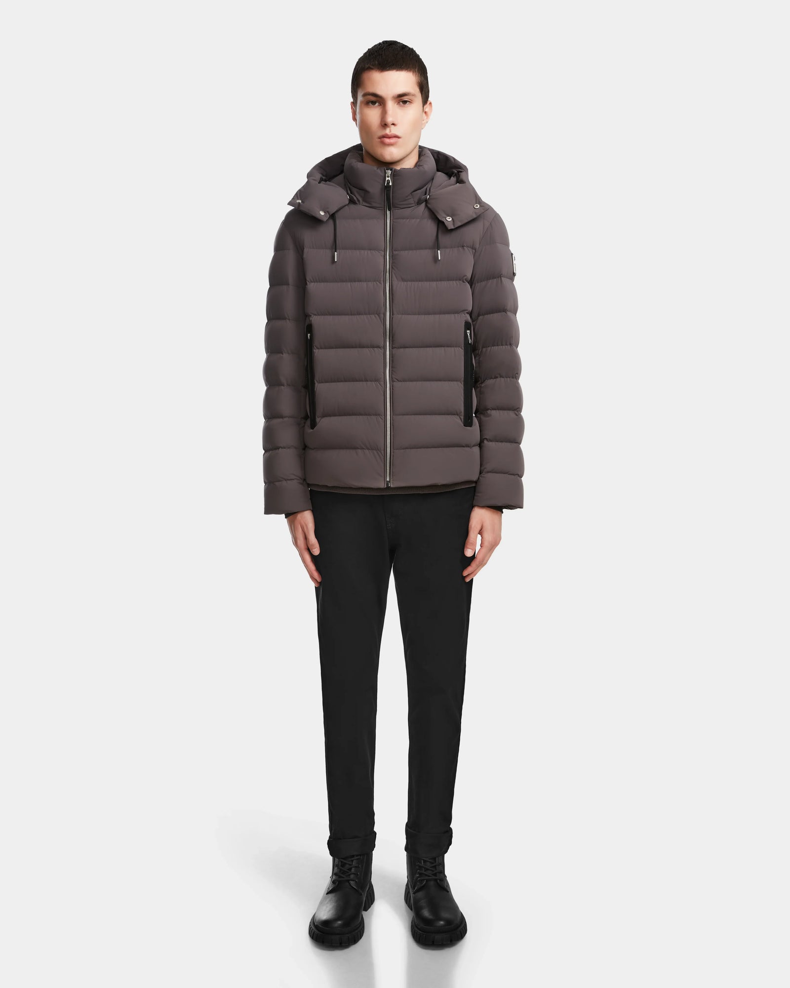 A person stands facing forward, wearing the Rudsak NAVAN - 6124551 Grey men’s quilted down puffer jacket with a hood. The wind and water-repellent, RDS-certified down jacket is set against a plain white background.
