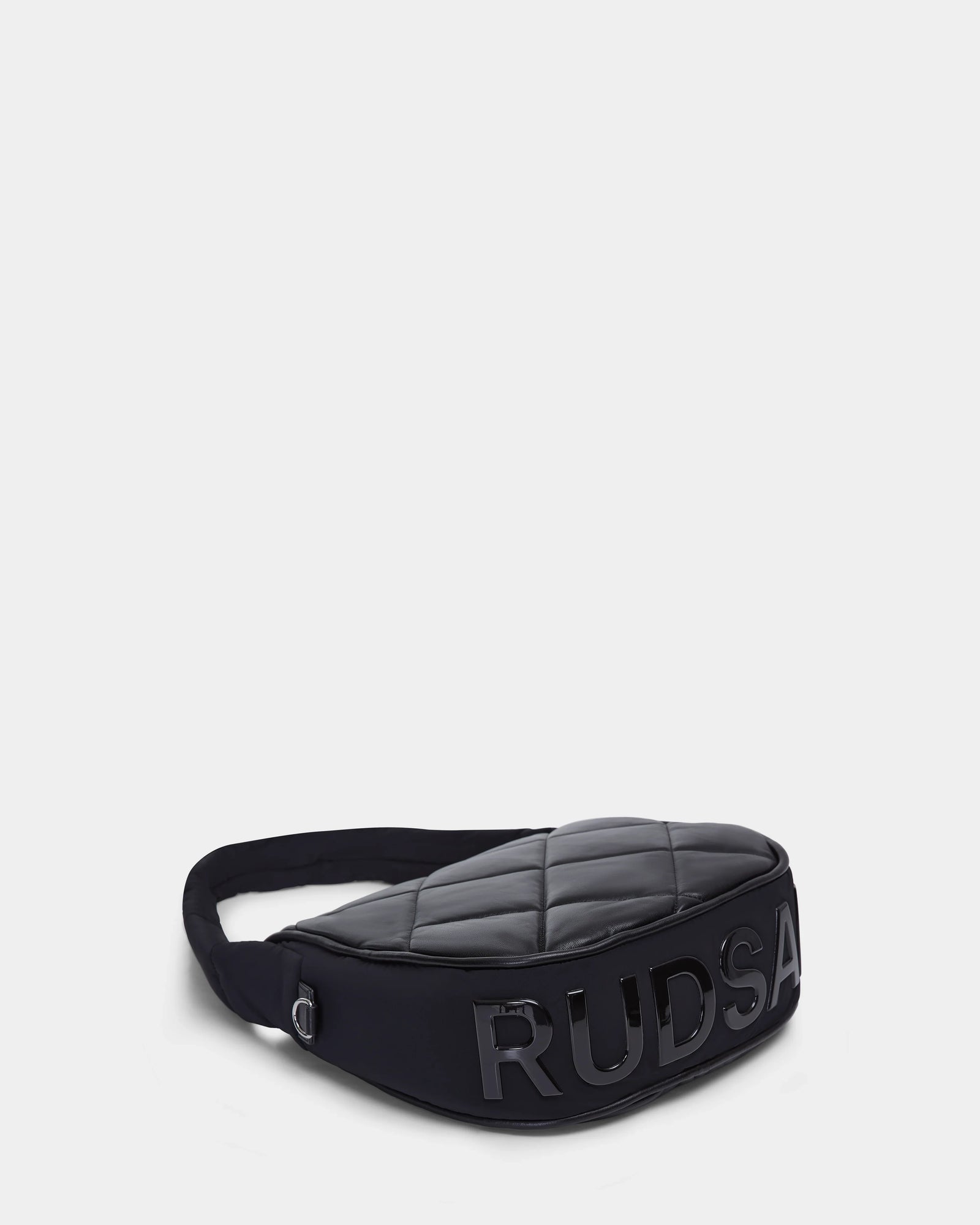 The NIOBE L - 8324503 Black by Rudsak is a quilted black crossbody bag with an adjustable strap and bold shiny black RUDSAK lettering on the front, shown against a plain white background.