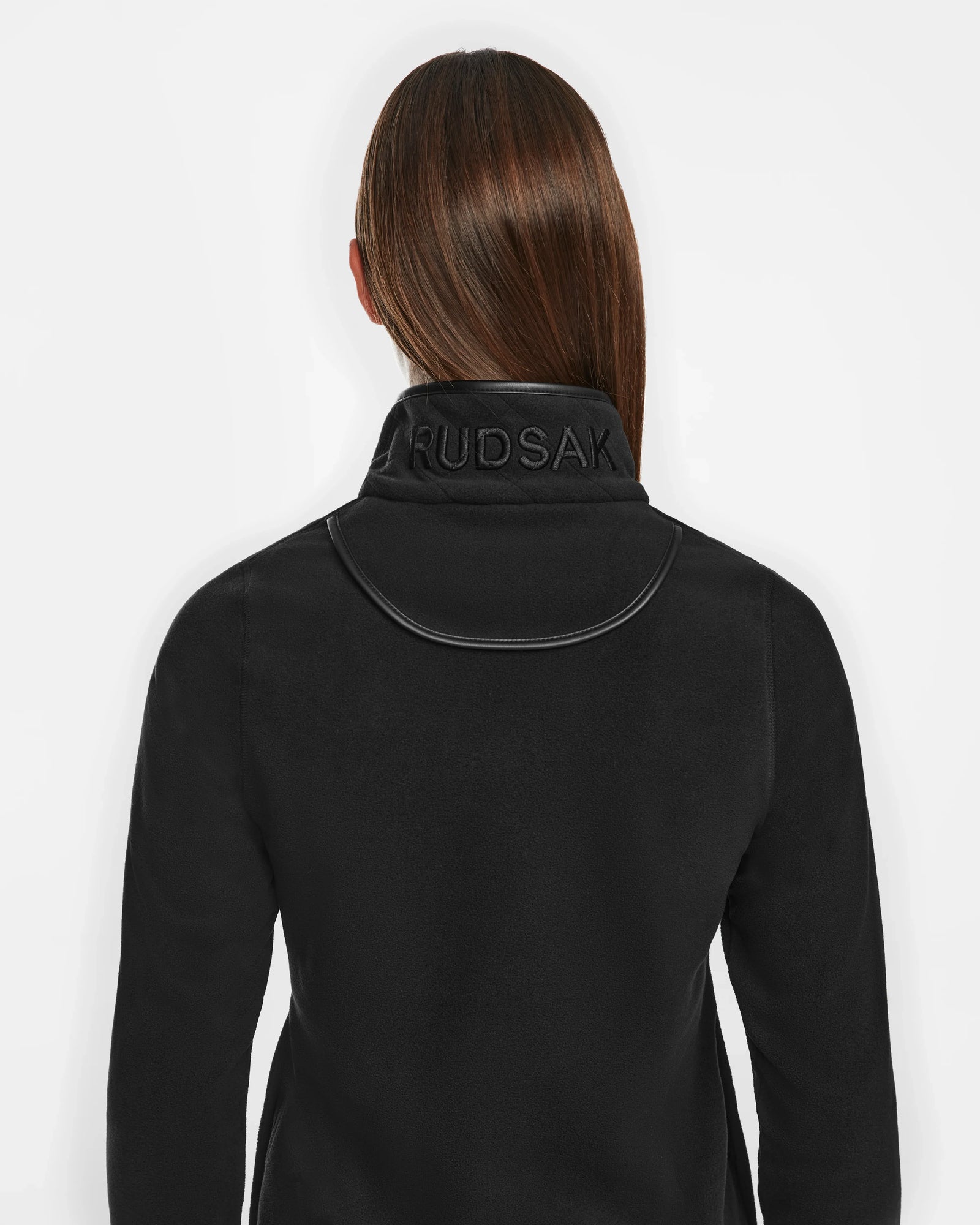 A person with straight brown hair wears the Rudsak ORCHID - 8524616 Black jacket, featuring vegan leather trims and a high collar with RUDSAK embossed on the back. The person faces away, against a white background.