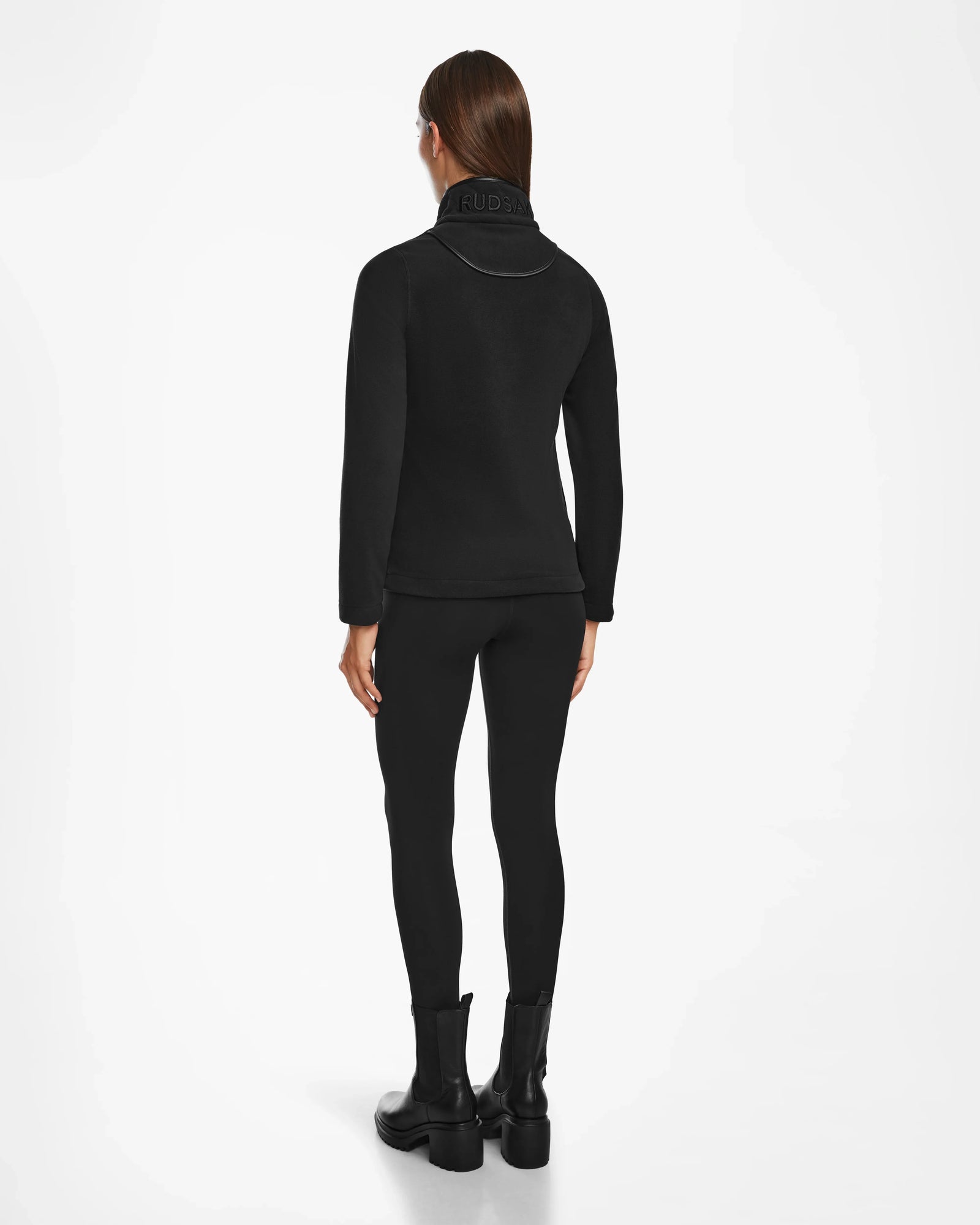 A person with straight brown hair models the Rudsak ORCHID - 8524616 Black jacket, featuring vegan leather trims, paired with black leggings and boots, standing facing away on a plain white background.