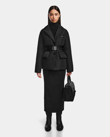 A woman with straight dark hair and a neutral expression stands against a white background, wearing the Rudsak PAOLA - 8124547 Black womens down blazer over a black turtleneck dress, black boots, and holding a black handbag.