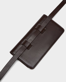 The Rudsak PREYA - 8324508 Espresso crossbody bag features dark brown leather, a long wide strap, smooth finish, and minimalist design, shown here against a light gray background.