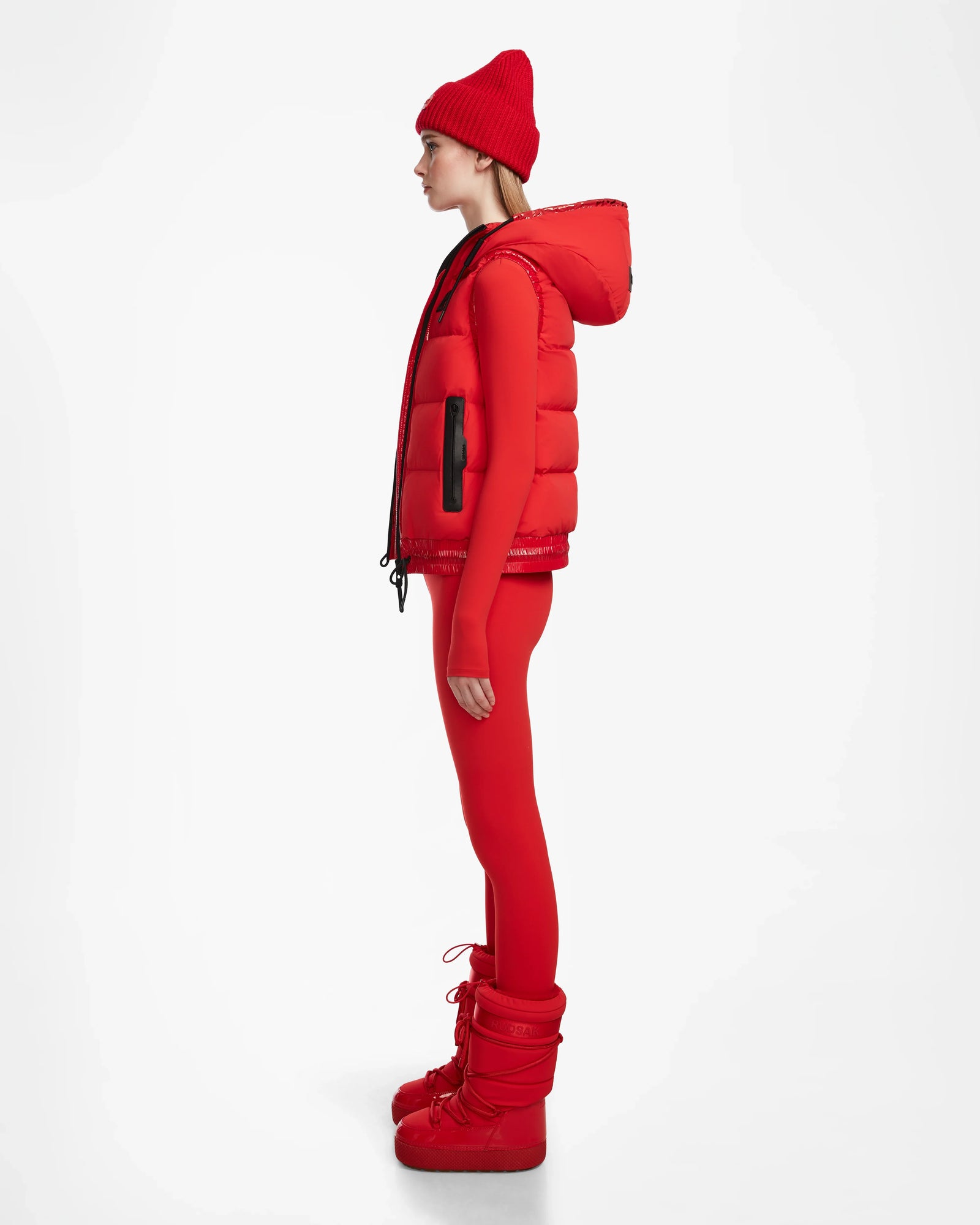 A person in profile wears the Rudsak RAQUEL M - 8124538 Flame Red reversible ski vest with hood, paired with matching red fitted leggings and padded boots, set against a plain white background.