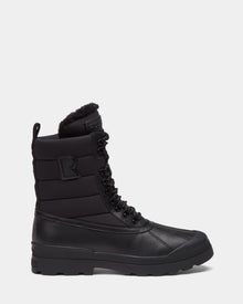 The Rudsak REAGAN - 6224501 Black is a mens black lace-up winter boot with a rugged sole, quilted upper, faux fur lining, side pull tab, embossed R logo, and high ankle design for warmth and style all season.