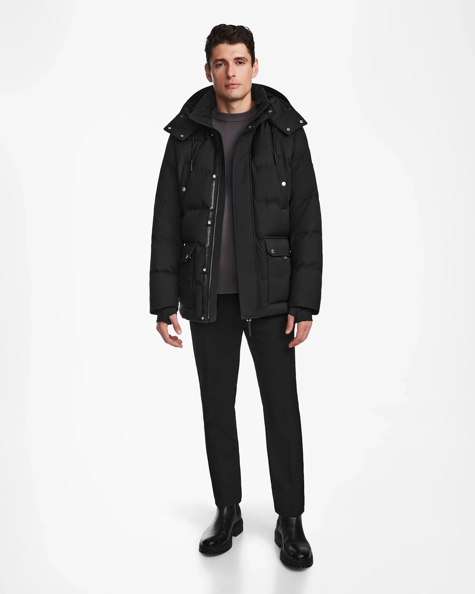 A man faces forward against a white background, wearing the Rudsak RICKY - M123502 Black parka over a dark sweater, black pants, and black boots. His posture is relaxed and his expression neutral.