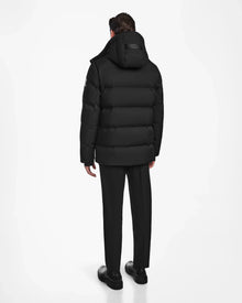 A person stands facing away, wearing the Rudsak RICKY - M123502 Black parka with a hood, paired with black pants and shoes, against a plain white background.