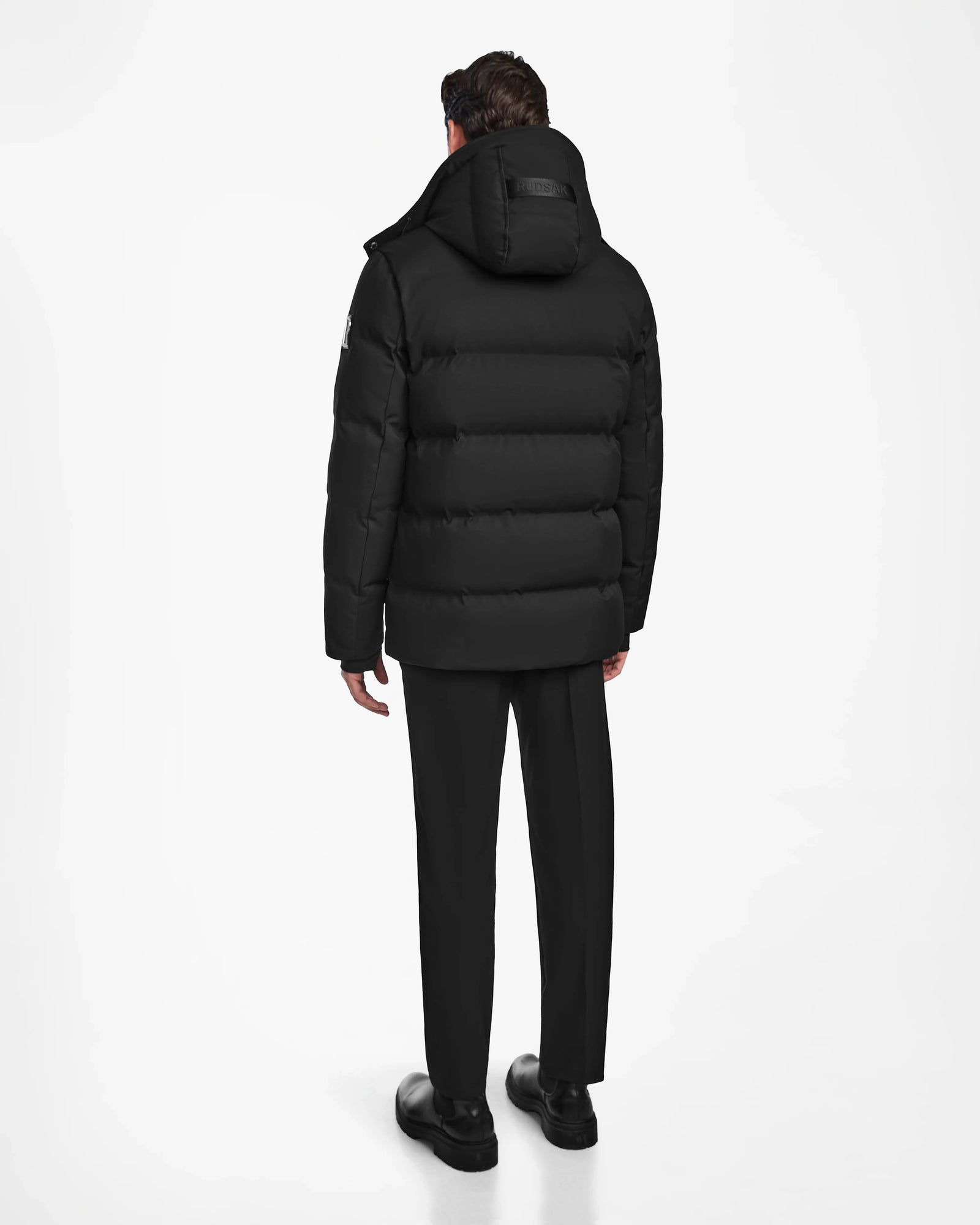 A person stands facing away, wearing the Rudsak RICKY - M123502 Black parka with a hood, paired with black pants and shoes, against a plain white background.