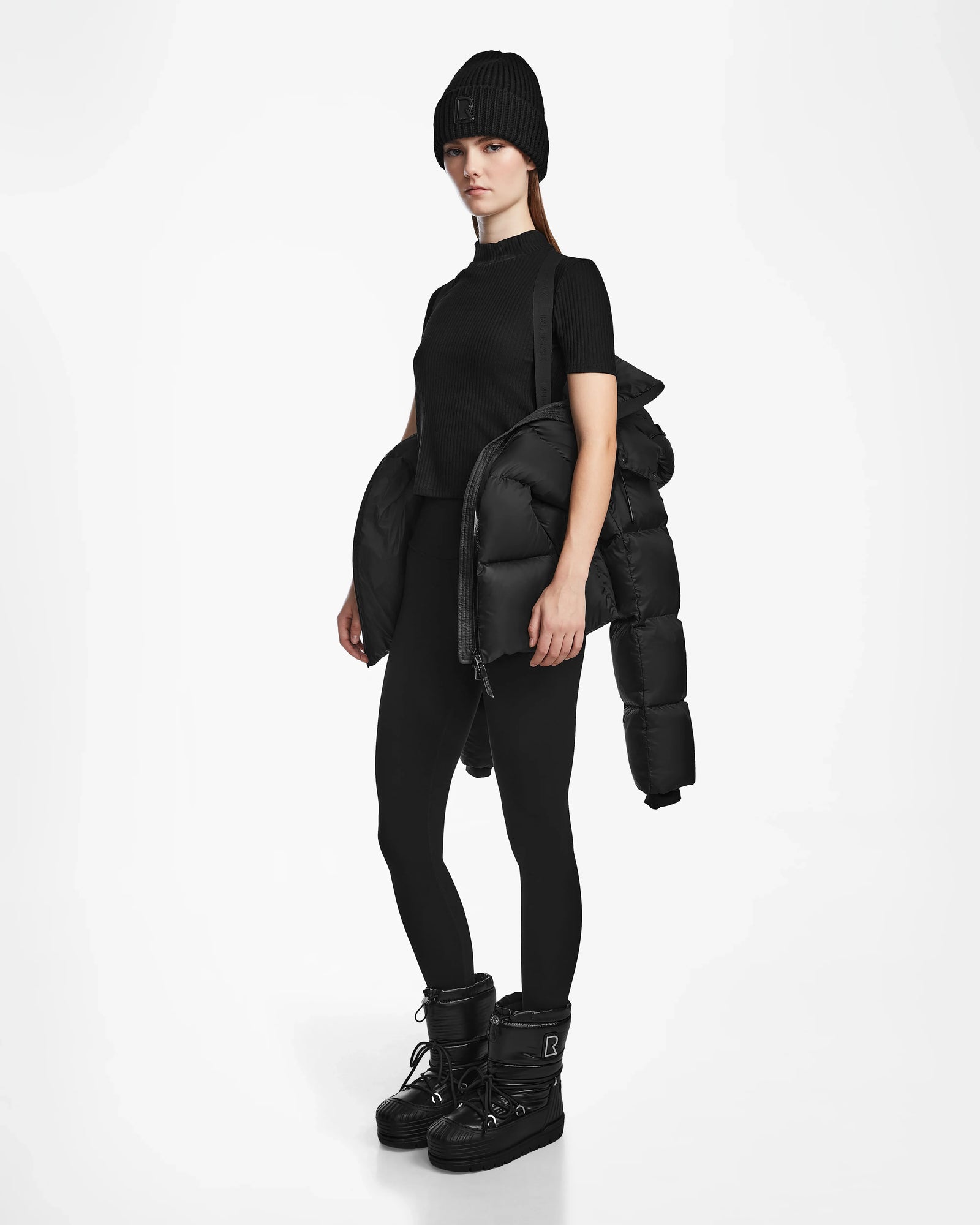 A woman poses against a white background wearing Rudsak's RITA M - L124537 Black weather-resistant puffer jacket draped over one shoulder, styled with a black beanie, short-sleeve top, leggings, and winter boots.