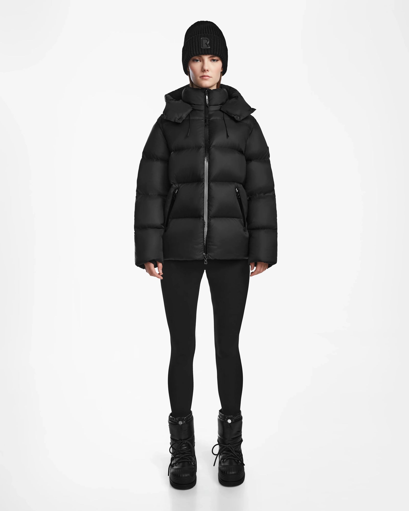 A person faces forward against a plain white background, wearing Rudsak's RITA M - L124537 Black weather-resistant puffer jacket, paired with black leggings, black winter boots, and a black knit beanie.