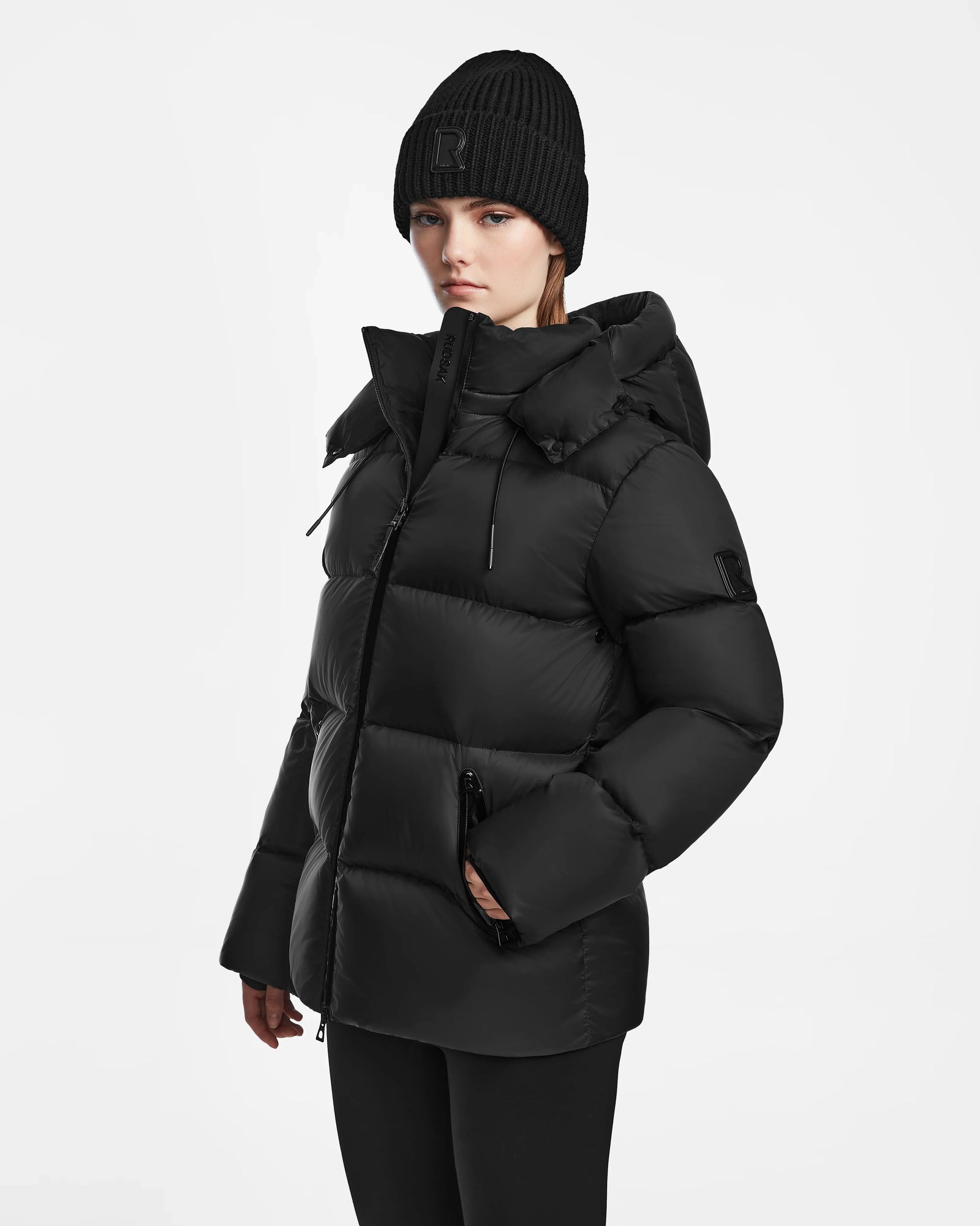 A woman in the Rudsak RITA M - L124537 Black weather-resistant hooded down puffer and a black knit beanie stands slightly turned against a plain white background, hands tucked into her coat pockets.