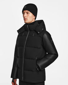 A man wearing the Rudsak RONNY M - M124513 Black puffer jacket with a hood and a black knit beanie stands against a plain white background, facing slightly left with a neutral expression.