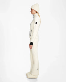 A woman stands in profile against a plain white background, dressed in an all-white outfit featuring Rudsak's ROSALIE - 8124784 Iceland women's fitted ski jacket, paired with a beanie, flared pants, and platform shoes.