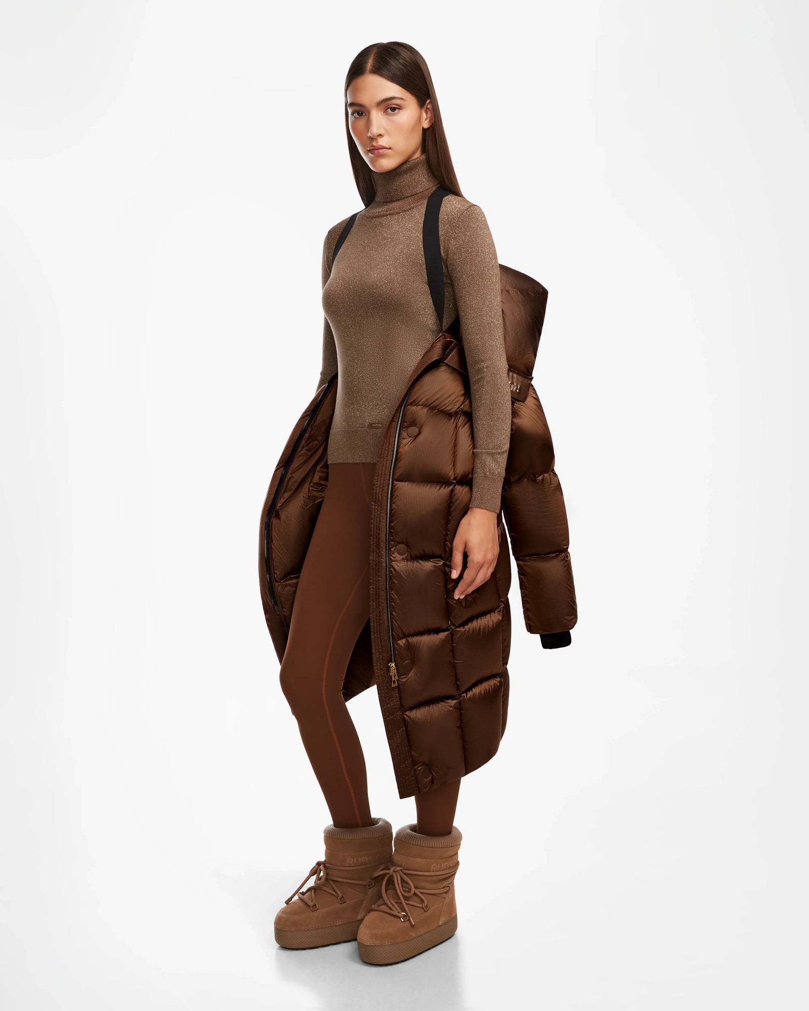 A woman poses with a neutral expression against a white background, draped in the Rudsak SASH R - 8124543 Antique Bronze RDS-certified down puffer jacket over brown leggings, a brown turtleneck, and brown puffer boots.