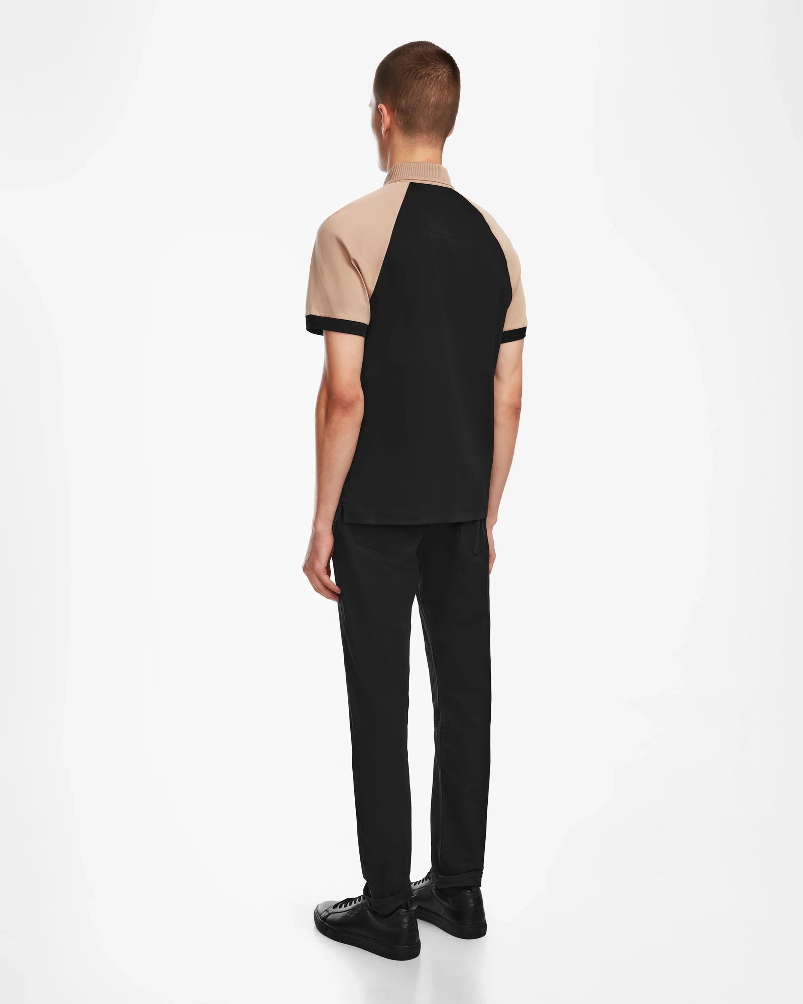 A man stands with his back to the camera, wearing the Rudsak WALTER - 6524531 Black/Camel premium cotton short sleeve polo, black pants, and black shoes against a plain white background.