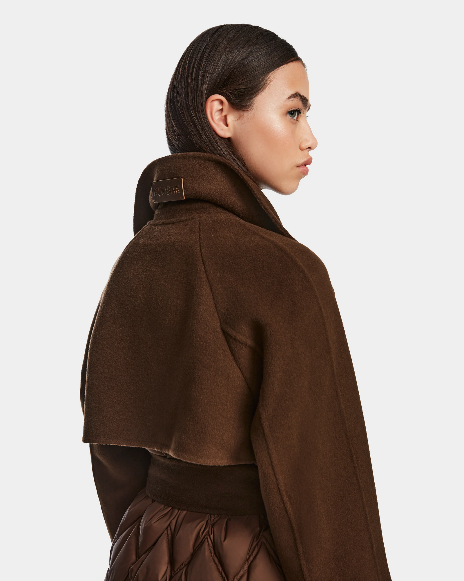 A woman in profile models the Rudsak ZOE - 8124711 Mocha, a stylish dark brown fringe belt coat with a wide collar, paired with a quilted brown skirt, set against a plain light background.