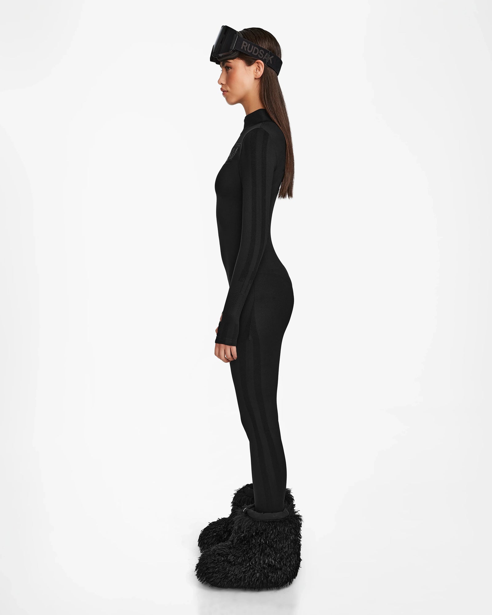 Wearing the Rudsak ZADIE - 8524609 Black long sleeve bodysuit, paired with fuzzy black boots and ski goggles, a person stands in profile against a plain white background.