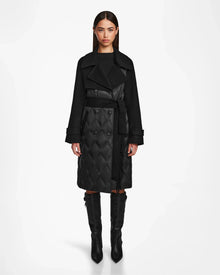 A woman faces forward in the Rudsak ZOE - 8124711 Black quilted down trench coat with a belted waist and tall black boots, set against a plain white background.