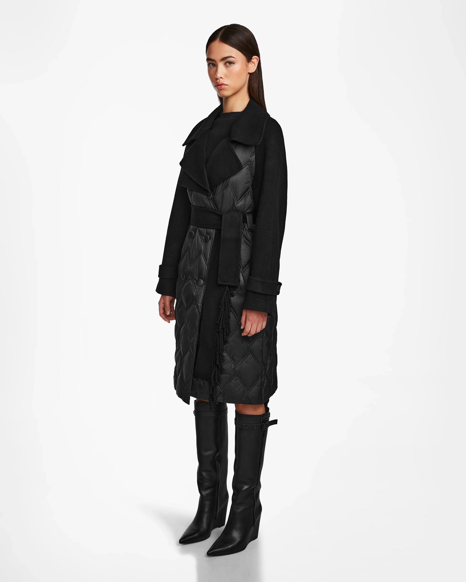 A woman with straight brown hair and a neutral expression models the Rudsak ZOE - 8124711 Black womens long quilted down trench coat with a wide collar and belted waist, paired with knee-high black heeled boots, against a white background.