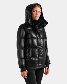 A woman wearing the Rudsak ALESSANDRA S-S - L125560 Black down puffer with a hood stands against a light gray background, gazing slightly to the side with a neutral expression.