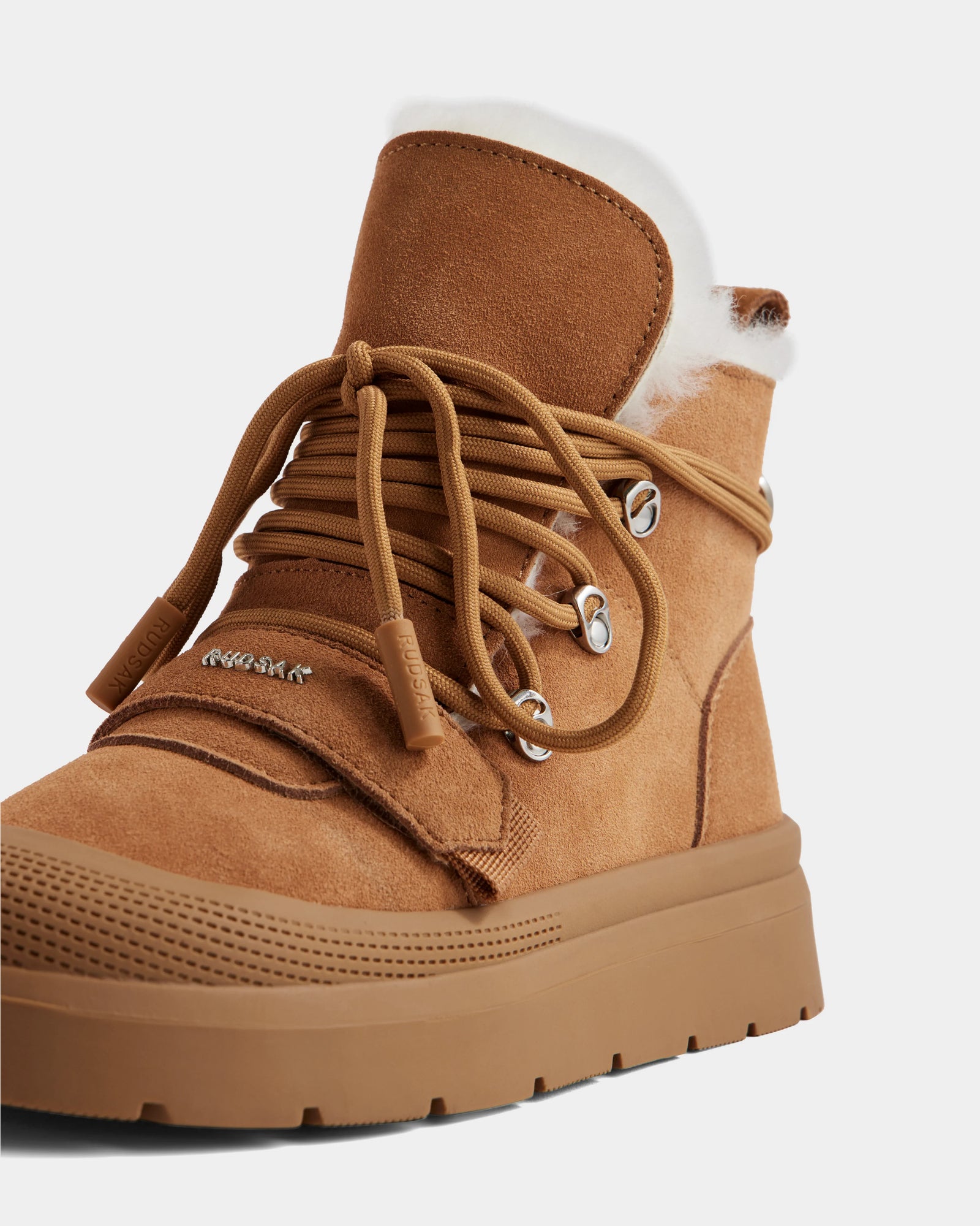 A close-up of Rudsak's ALEXEY - L225025 Tan women's suede winter boot, featuring thick brown laces, metal eyelets, a chunky rubber sole, and a soft wool lining visible at the top for extra warmth and comfort.
