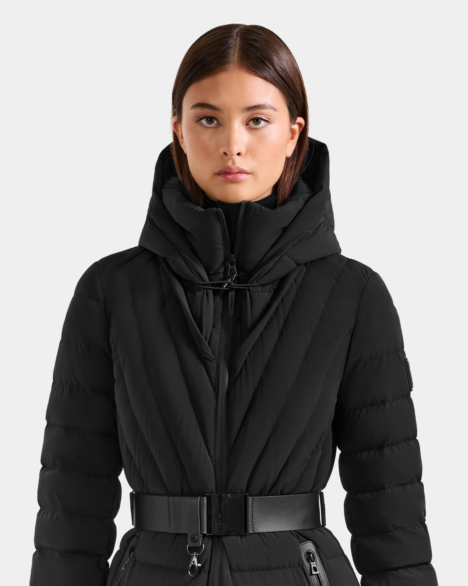 A woman with straight brown hair wears the Rudsak ALISA L - L125574 Black belted, hooded quilted puffer jacket. She faces forward against a plain light gray background, looking directly at the camera.