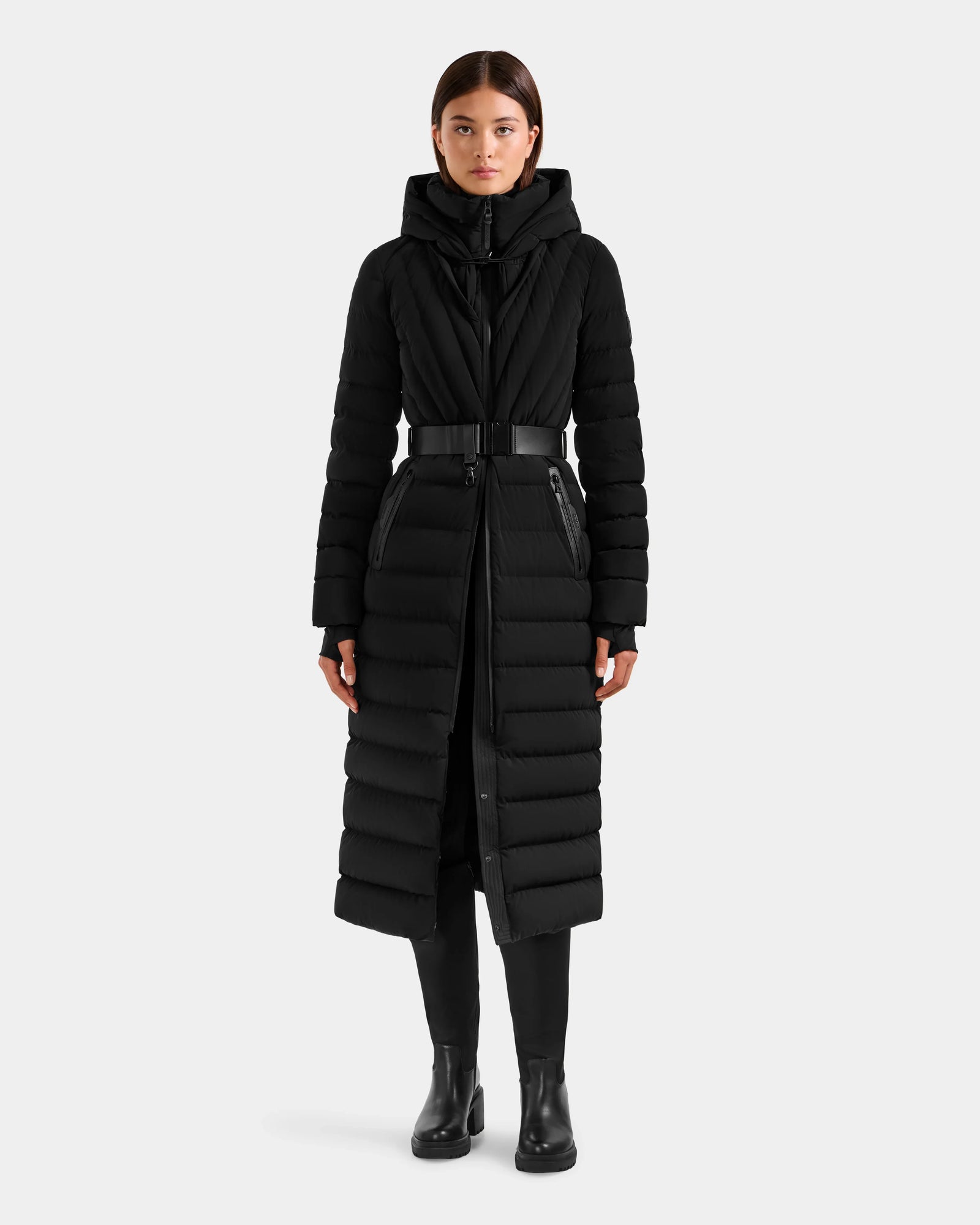 A woman faces forward in a Rudsak ALISA L - L125574 Black quilted puffer coat with hood, faux-fur trim, waist belt, and zippered pockets, paired with black pants and ankle boots against a plain light background.