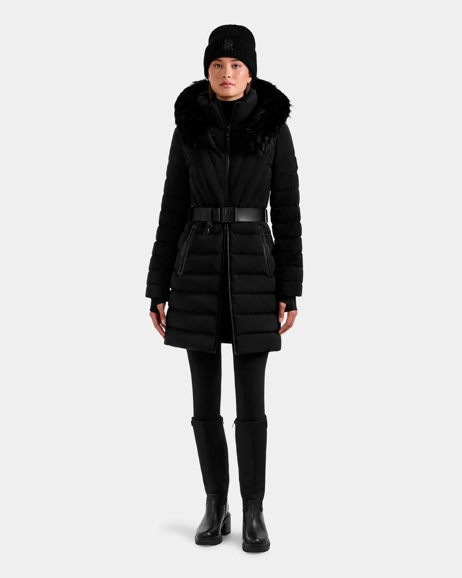 A person wearing the Rudsak ALISA M - L125586 Black fitted quilted down parka with fur-trimmed hood, black pants, knee-high boots, and a black beanie stands against a plain light background.
