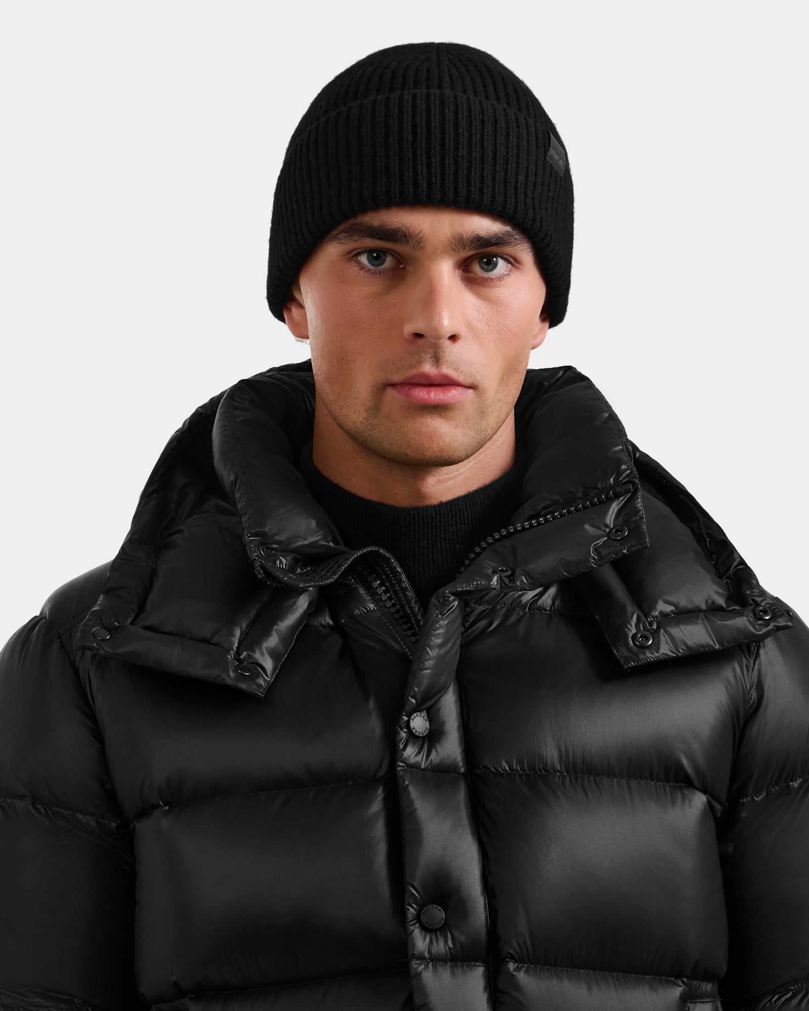 Wearing the Rudsak ANDREY - U425507 Black puffer jacket and a rib-knit beanie, a person stands against a plain light background, looking directly at the camera with a neutral expression.