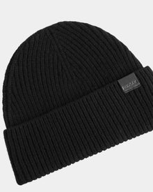The ANDREY - U425507 Black by Rudsak is a unisex black ribbed knit beanie with a folded cuff, featuring a small rectangular Rudsak logo patch on the cuff for classic style and comfort.