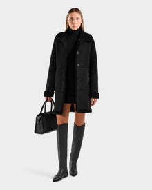 A woman stands against a plain light background wearing the Rudsak ANGILICA - L125600 Black faux shearling knee-length coat, styled with a black turtleneck dress, black knee-high boots, and holding a black handbag.
