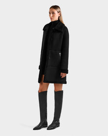 A woman wears the Rudsak ANGILICA - L125600 Black reversible knee-length coat with large front pockets, paired with a black turtleneck and knee-high boots, standing slightly to the side against a plain light background.