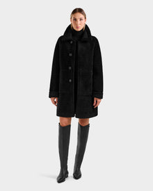 A woman faces forward against a light gray background, wearing the Rudsak ANGILICA - L125600 Black faux suede knee-length coat over a black turtleneck, paired with knee-high black boots.