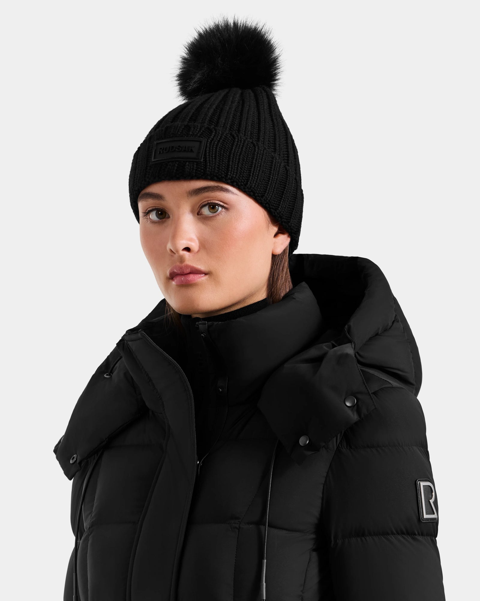 A woman wears the Rudsak ANISA - L423500 Black/Black padded winter jacket and a women's knit pom pom hat with faux fur, looking to the side against a plain light background—stylish essentials for any winter wardrobe.