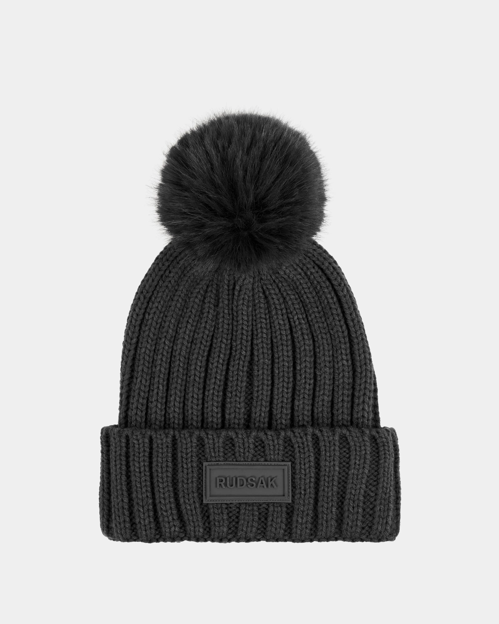 The ANISA - L423500 Grey by Rudsak is a ribbed knit beanie in grey with a large faux fur pom-pom, folded brim, and rectangular RUDSAK patch—an essential winter accessory. Shown against a plain light gray background.