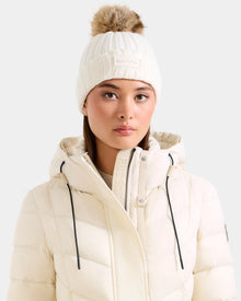 A woman models Rudsak’s ANISA - L423500 Optic White knit beanie with faux fur pom-pom and matching puffer jacket, both labeled Rudsak, against a light gray background—essential pieces for your winter wardrobe.