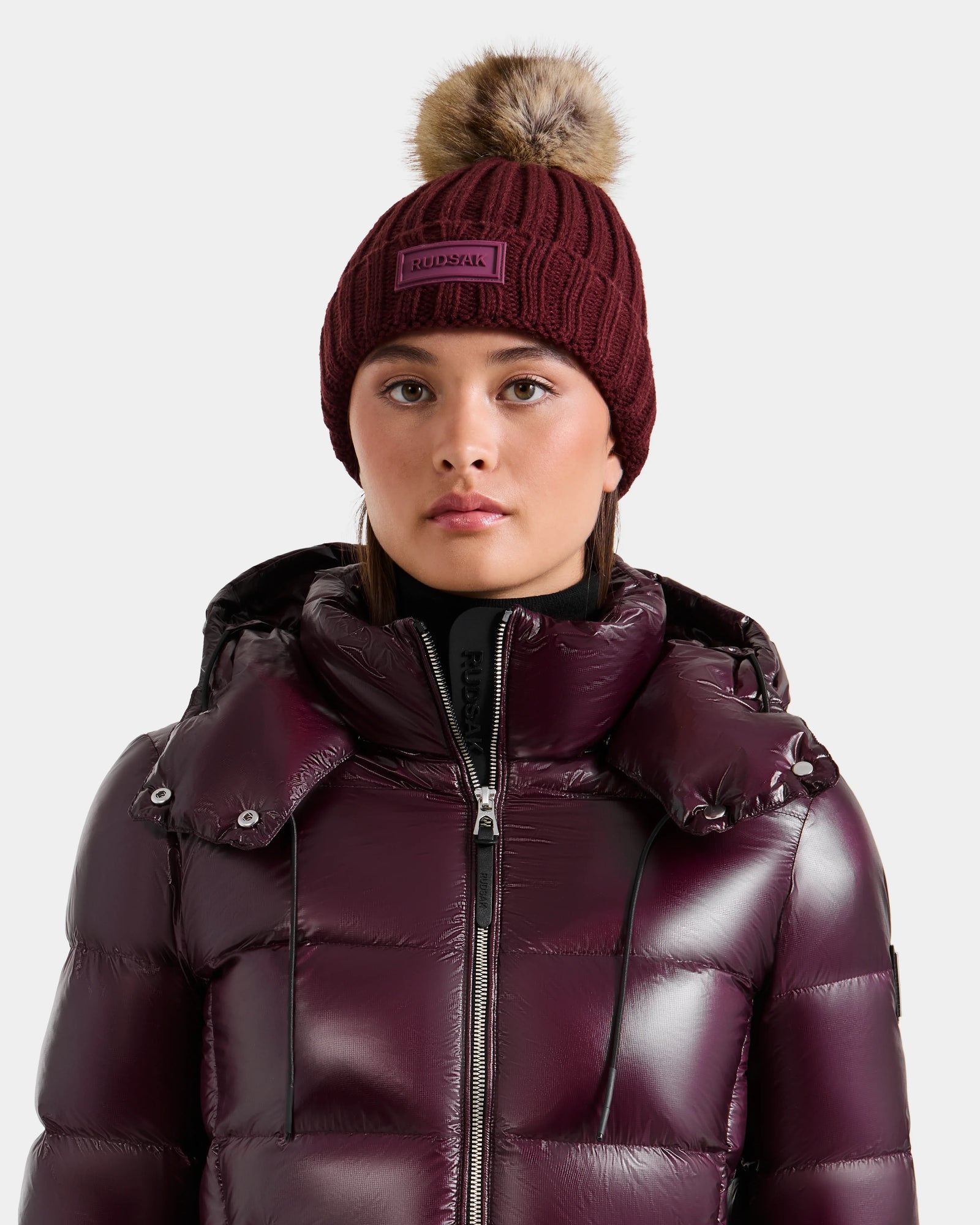 A person wearing the Rudsak ANISA - L423500 Wine puffer jacket and a matching maroon women's knit hat with faux fur pom-pom faces forward against a plain background, highlighting chic winter essentials.