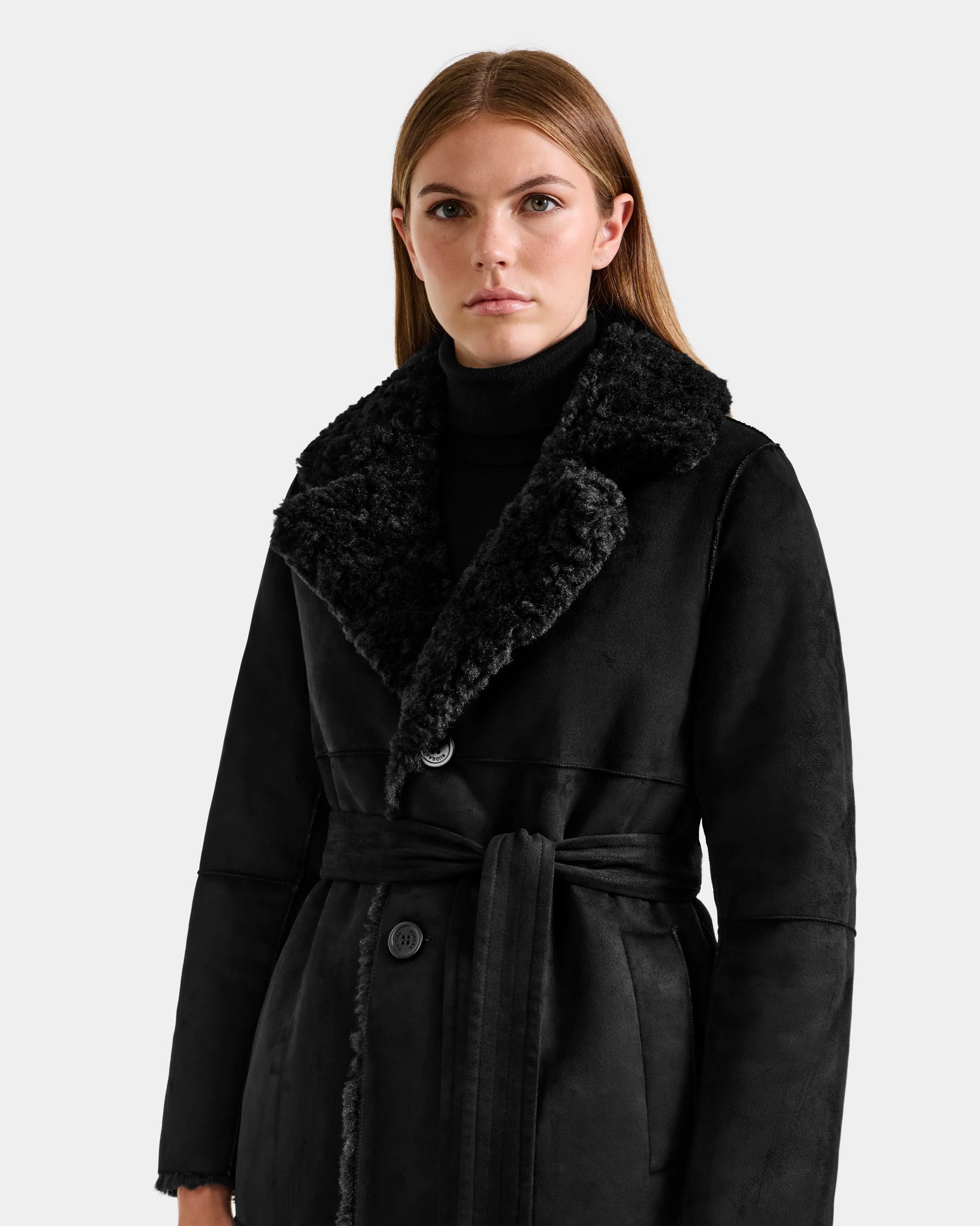 A woman with long brown hair models the Rudsak ANNABELLE FSU - L125512 Black faux suede shearling coat with a wide textured collar over a black turtleneck, standing against a plain light gray background.