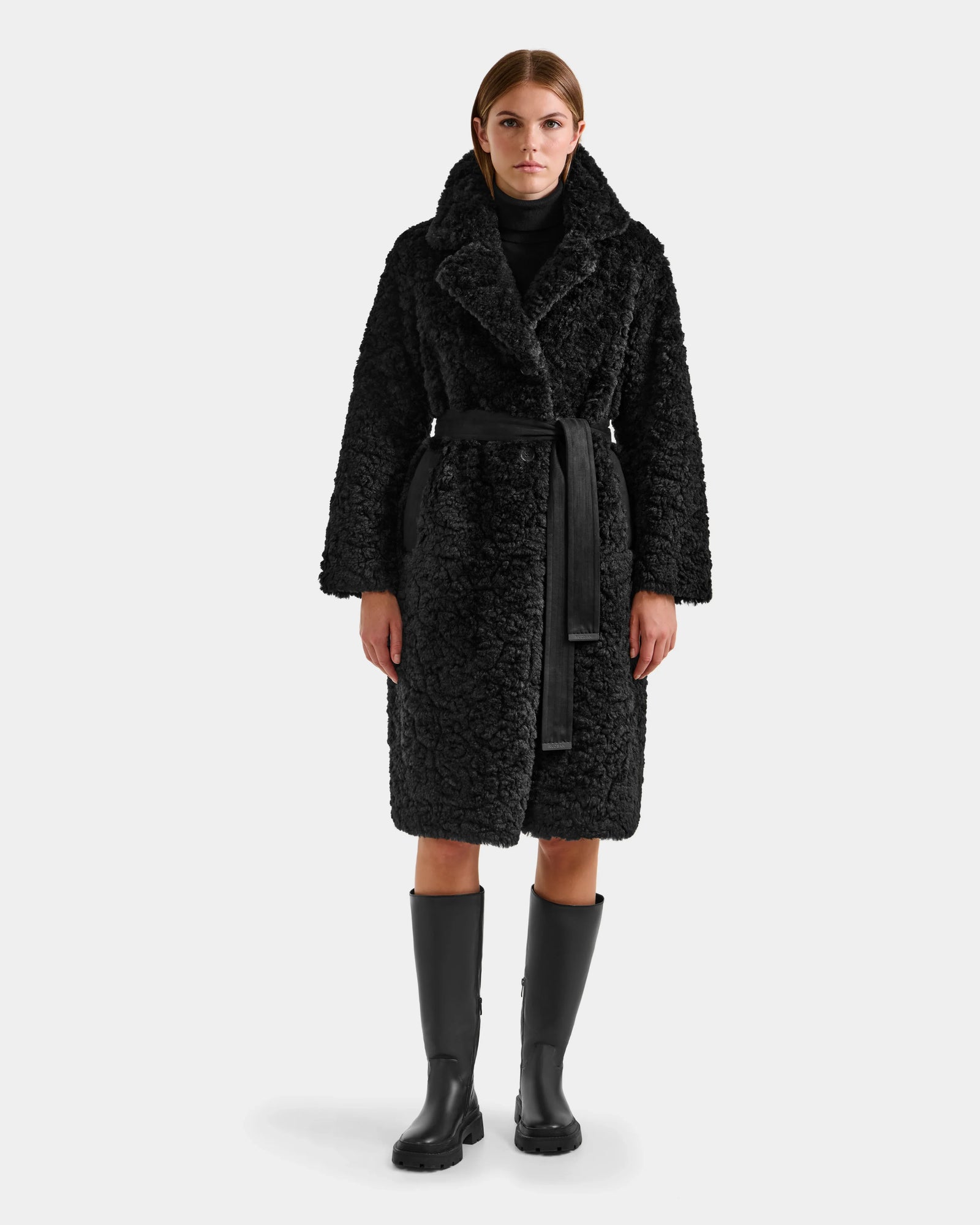 A woman faces forward in the Rudsak ANNABELLE FSU - L125512 Black teddy trench coat, black turtleneck, and black knee-high boots against a plain light gray background.