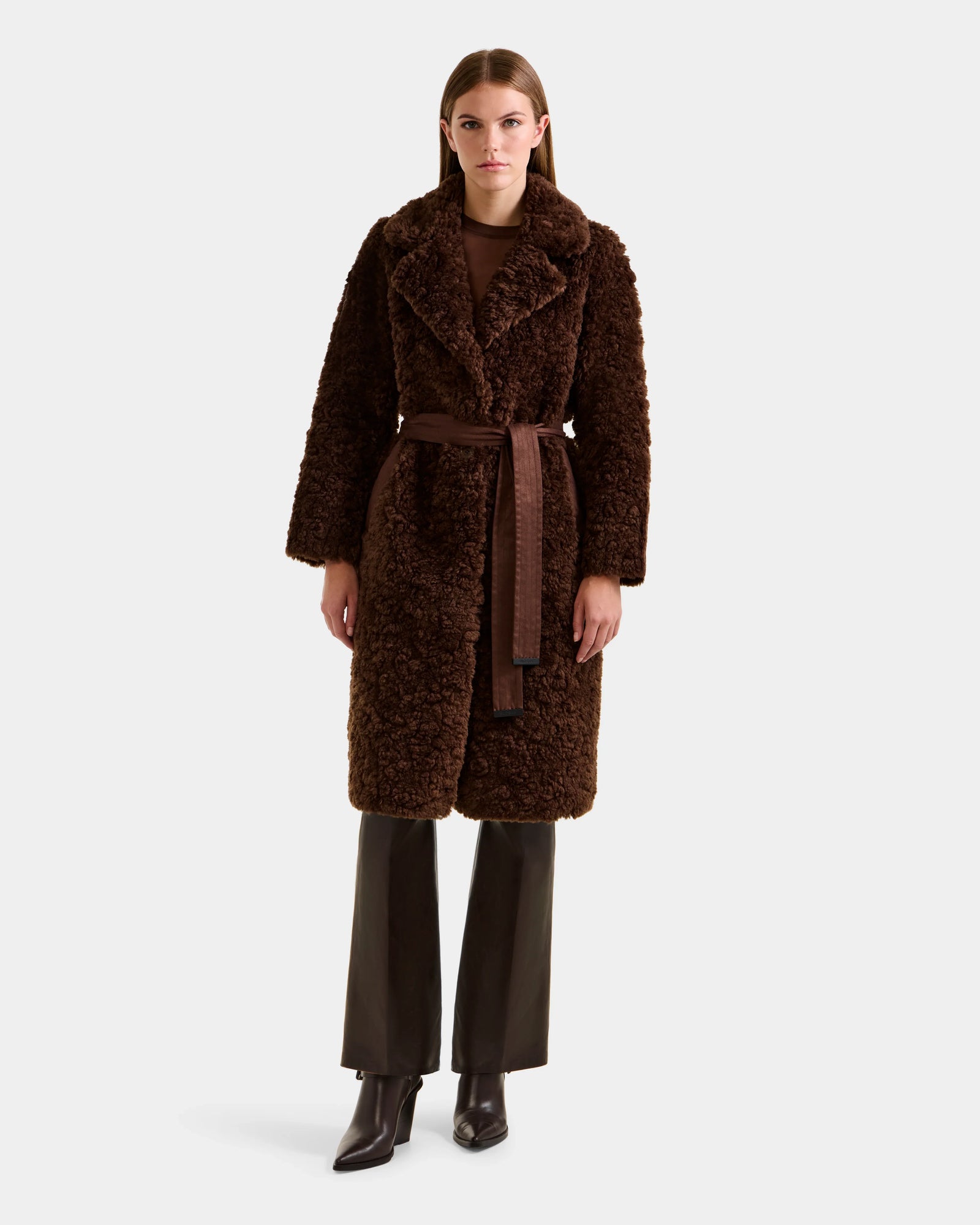 A woman stands facing forward in the Rudsak ANNABELLE FSU - L125512 Espresso, a long dark brown faux suede trench coat with matching belt, styled with brown pants, a brown top, and heeled boots against a plain light background.