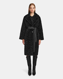 A woman stands facing forward, wearing the Rudsak ANNABELLE - L124544 black textured knee-length vegan leather coat with a wide collar and belted waist, paired with a black button-up top and knee-high black boots against a plain white background.