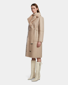 A woman wears the Rudsak ANNABELLE - L124544 Sand long vegan leather coat with a belt and wide collar, paired with knee-high cream boots, standing slightly turned against a plain white background.
