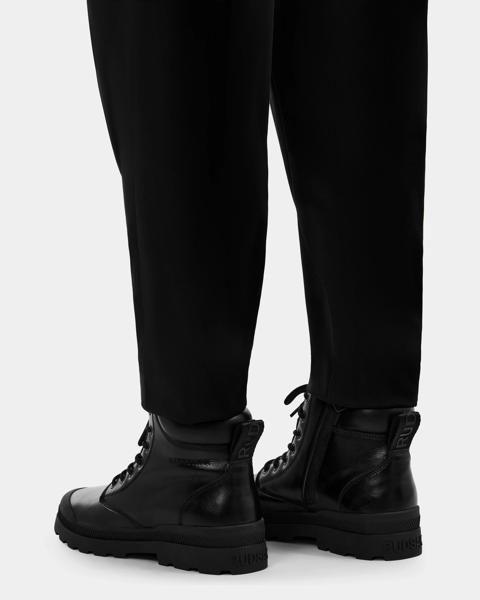 A person in black pants and Rudsak ARTUR - M223501 Black weather-treated men's ankle boots with thick soles, laces, side zippers, and heel branding stands on a light gray surface.