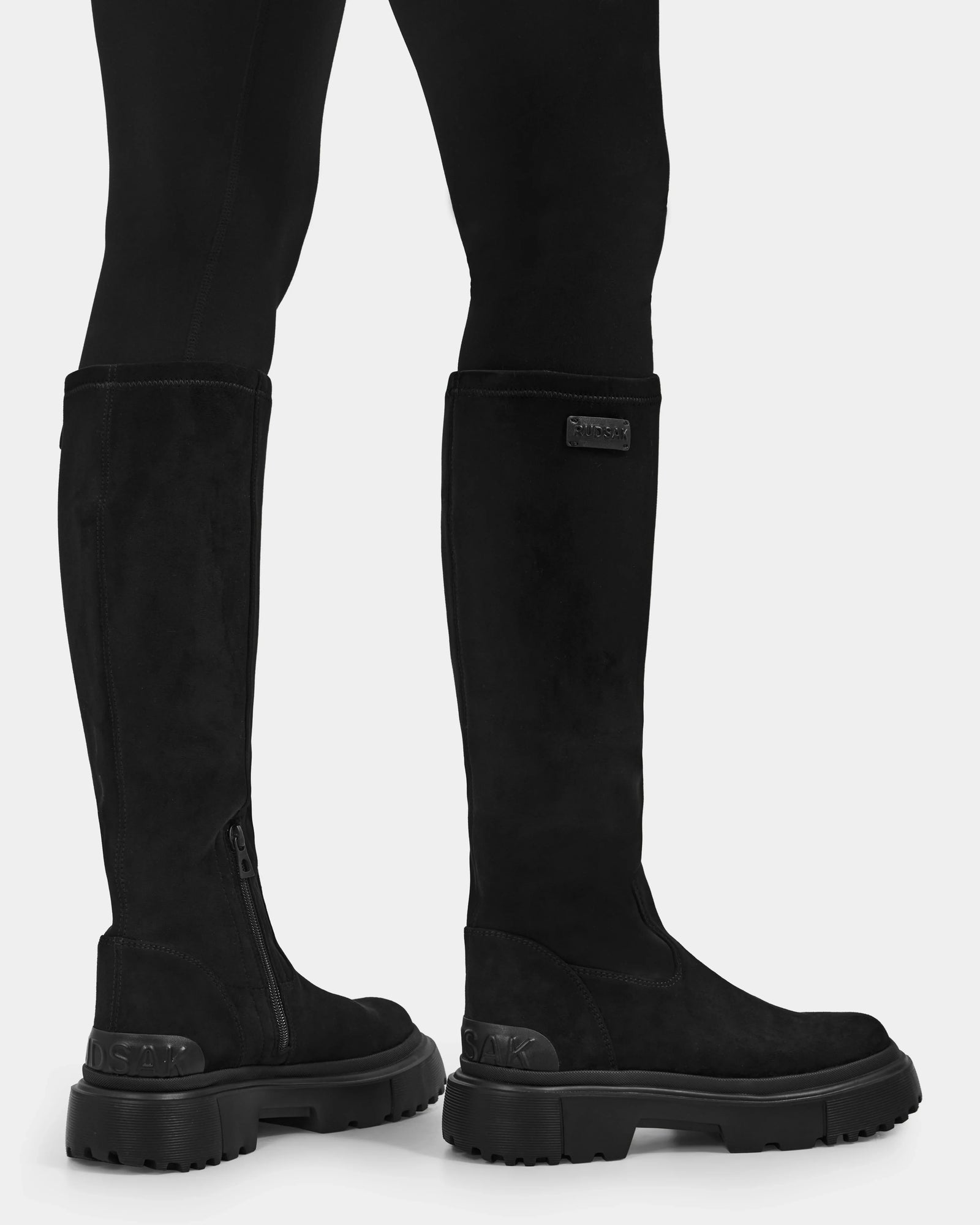 A person wears Rudsak AVOLA - L225514 Black women’s knee-high boots with chunky soles and side zippers, shown from the side and back against a white background. The boots are styled with black leggings for a sleek look.