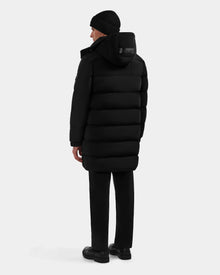 A person wearing the Rudsak BRAYDEN M-M - M125535 Black down puffer coat, black pants, shoes, and a beanie stands facing away against a plain light background, showcasing the coat’s winter versatility.