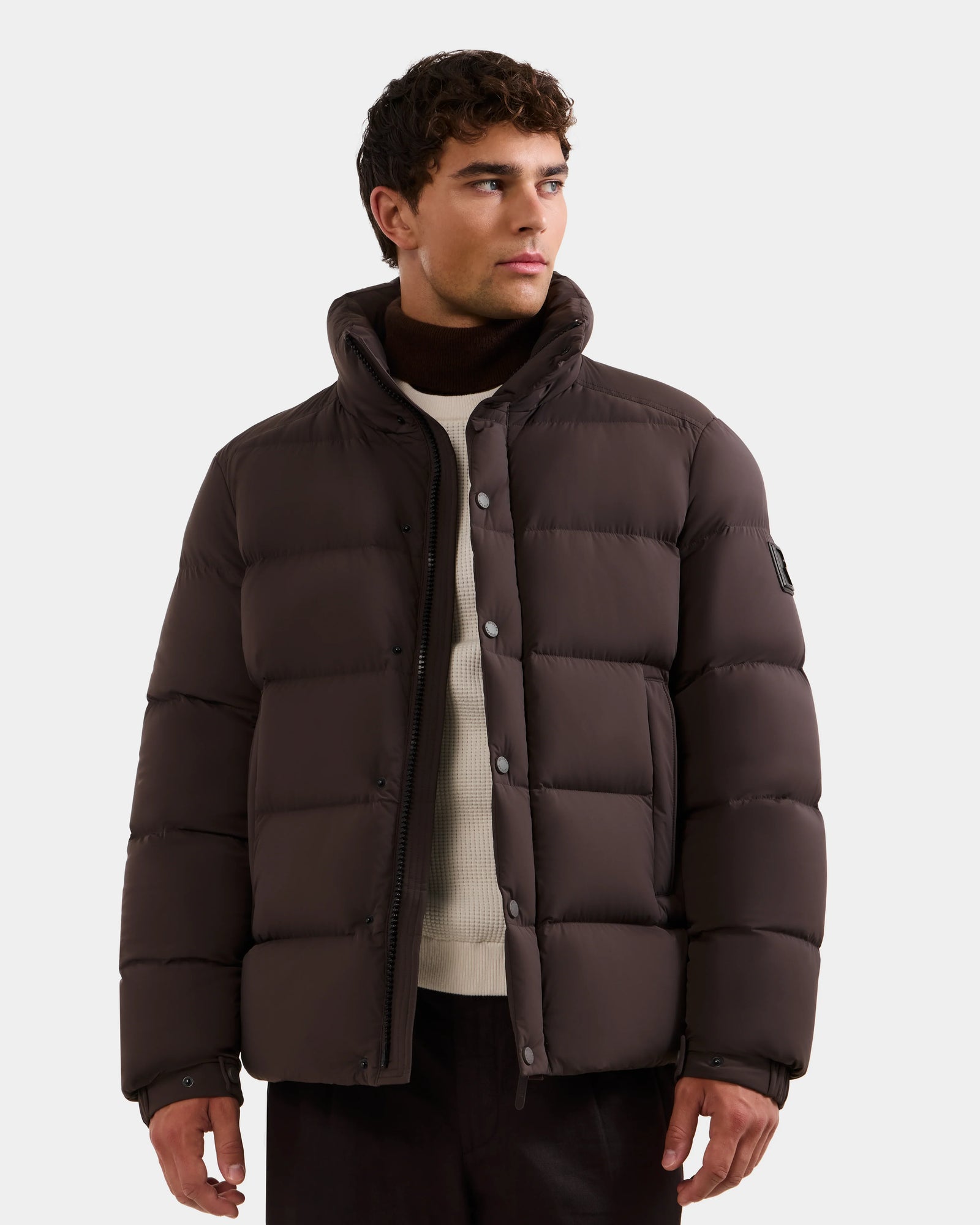 A man wearing the Rudsak BRAYDEN M-S - M125524 Espresso, an RDS-certified dark brown down puffer jacket with a high collar, stands against a plain light background over a cream sweater and dark pants, looking slightly to his right.