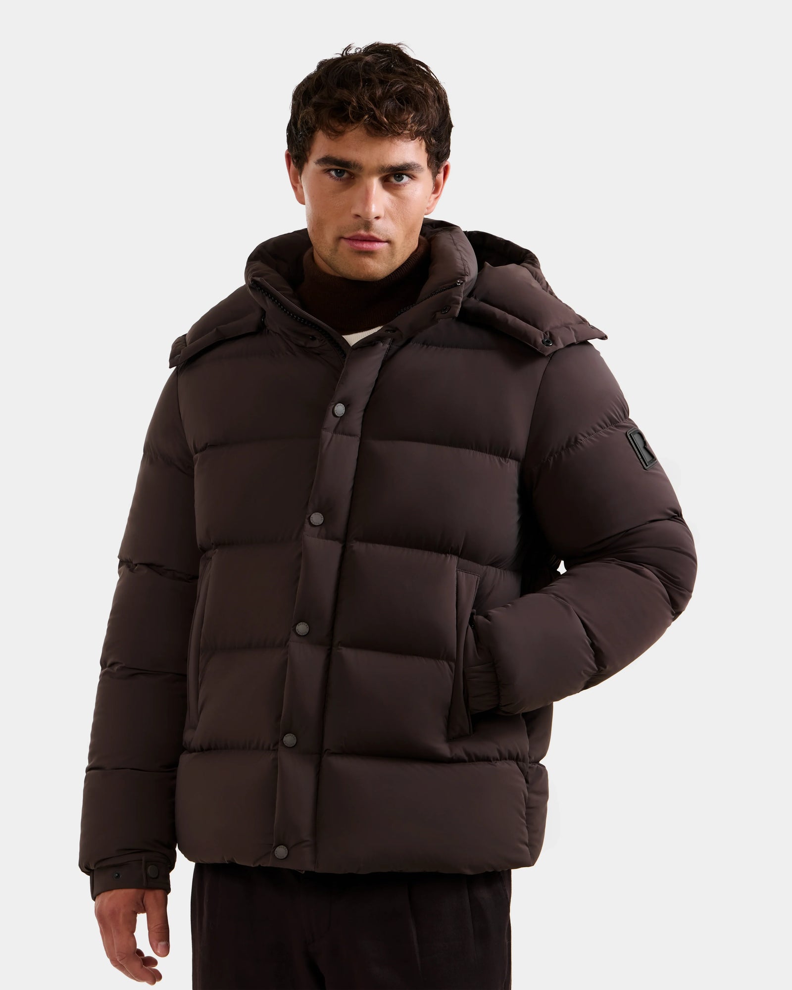 A man stands with one hand in his pocket against a plain background, wearing the Rudsak BRAYDEN M-S - M125524 Espresso down puffer jacket, featuring a hood and snap buttons.