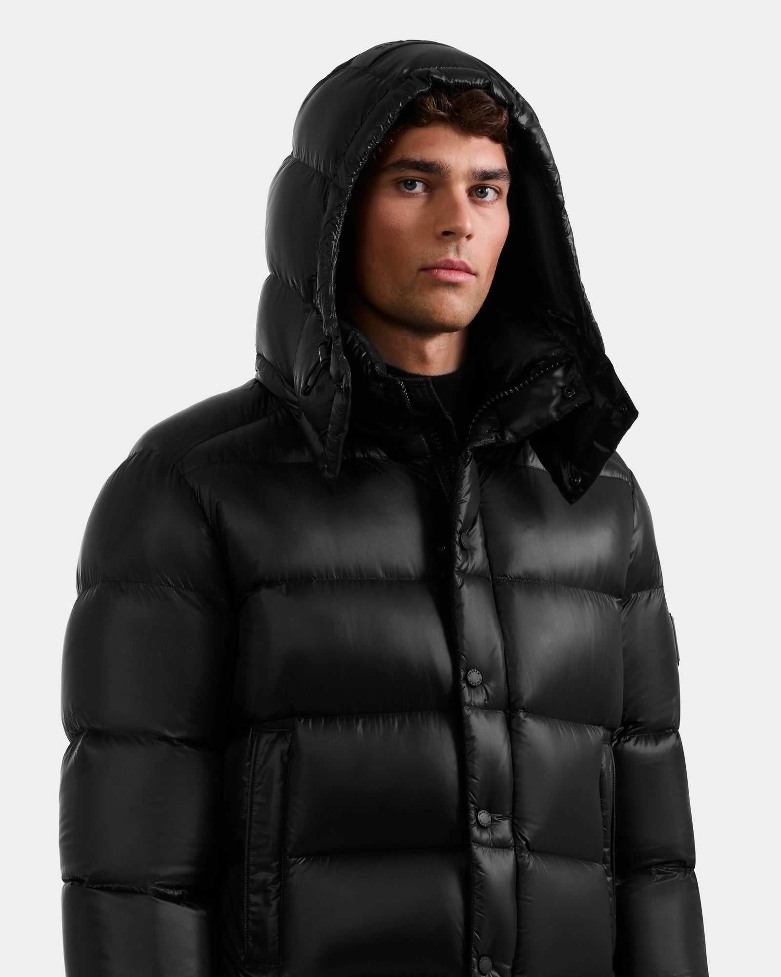 A young man wears the Rudsak BRAYDEN S - M125525 Black quilted down puffer jacket with the hood up, looking straight ahead against a plain light background.