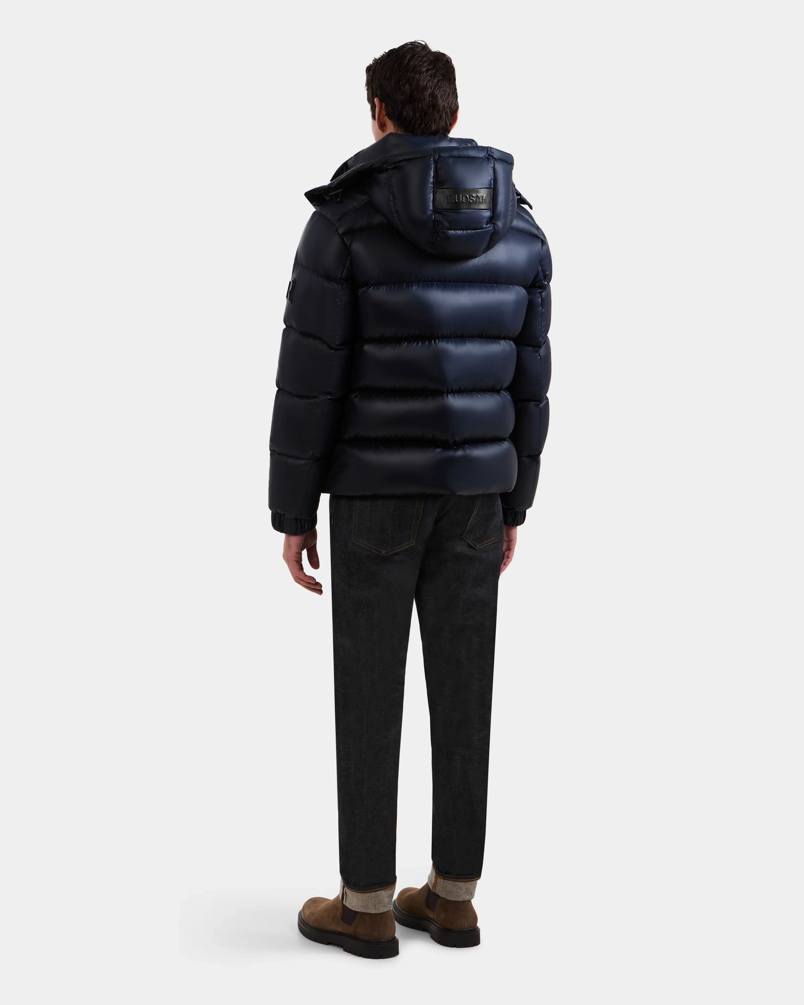 A man, seen from behind, wears the Rudsak BRAYDEN S - M125525 Navy shiny quilted down puffer with hood, paired with dark jeans and brown shoes, set against a plain light background.