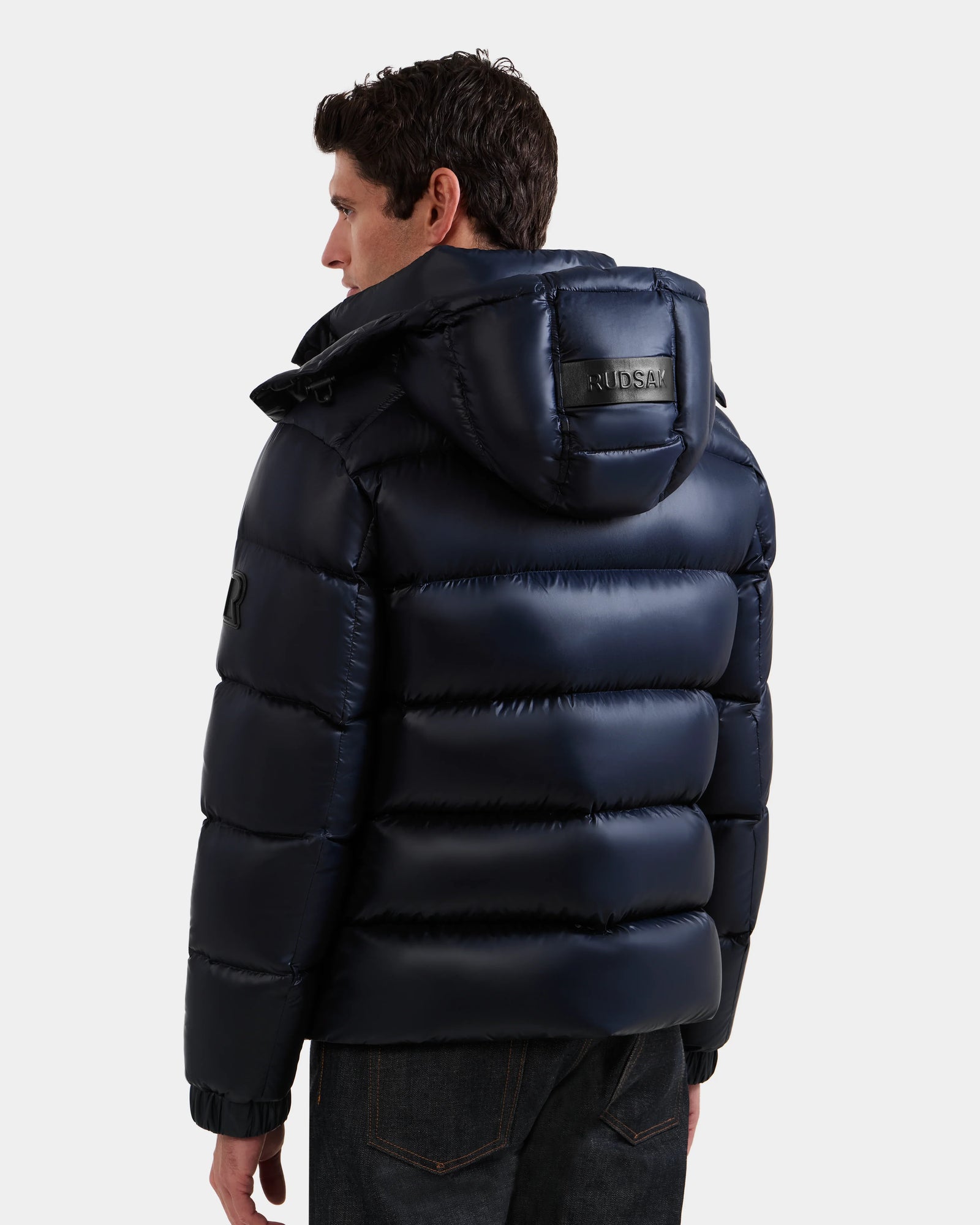 A man wearing the BRAYDEN S - M125525 Navy, a shiny dark navy men's winter down puffer jacket with a hood and visible stitching from Rudsak, stands facing away; the brand's patch is displayed on the hood against a light background.