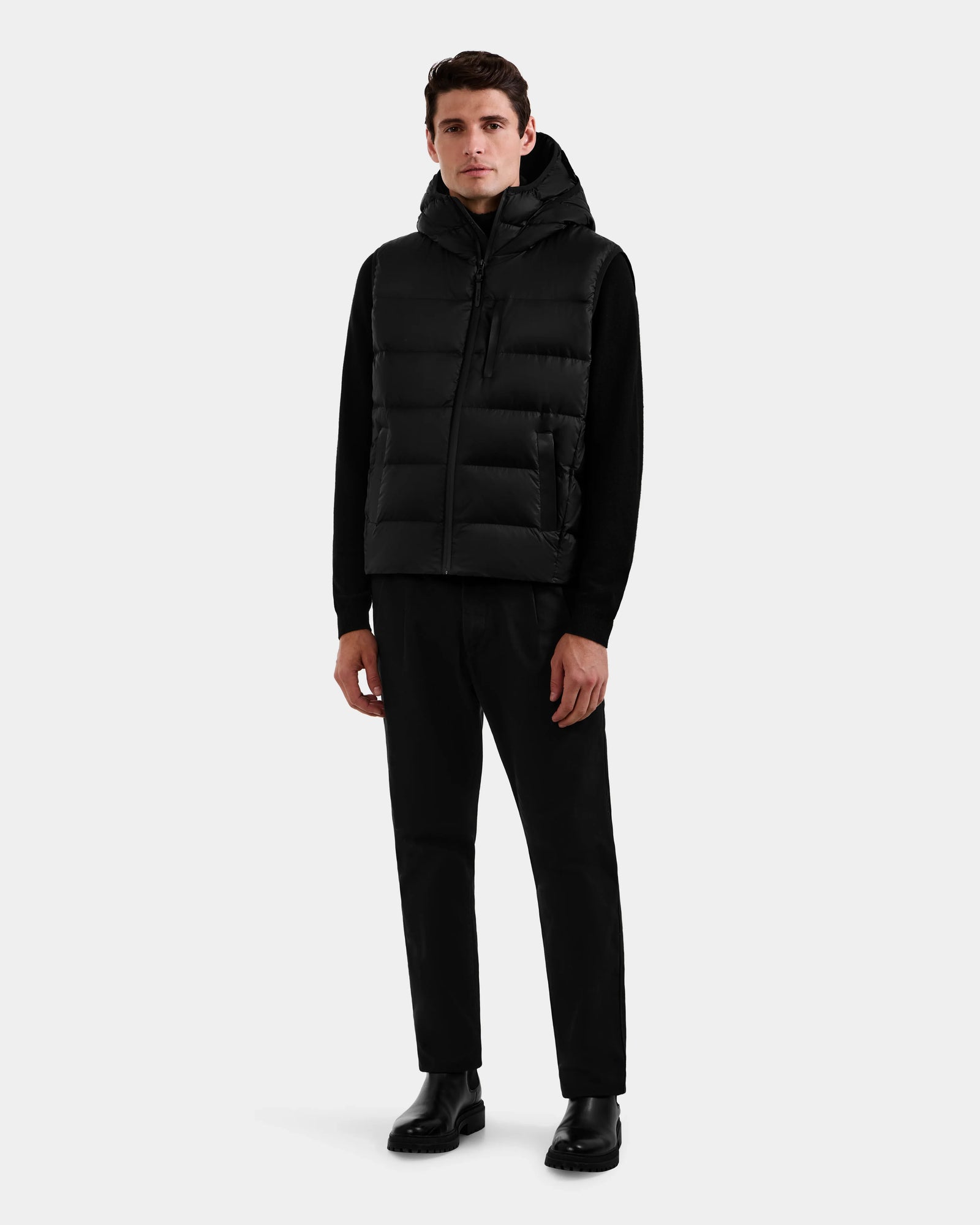 A man faces forward, wearing the Rudsak BROOKS - M125520 Black men’s down puffer vest over a black long-sleeve shirt, with black pants and boots, set against a plain light gray background.